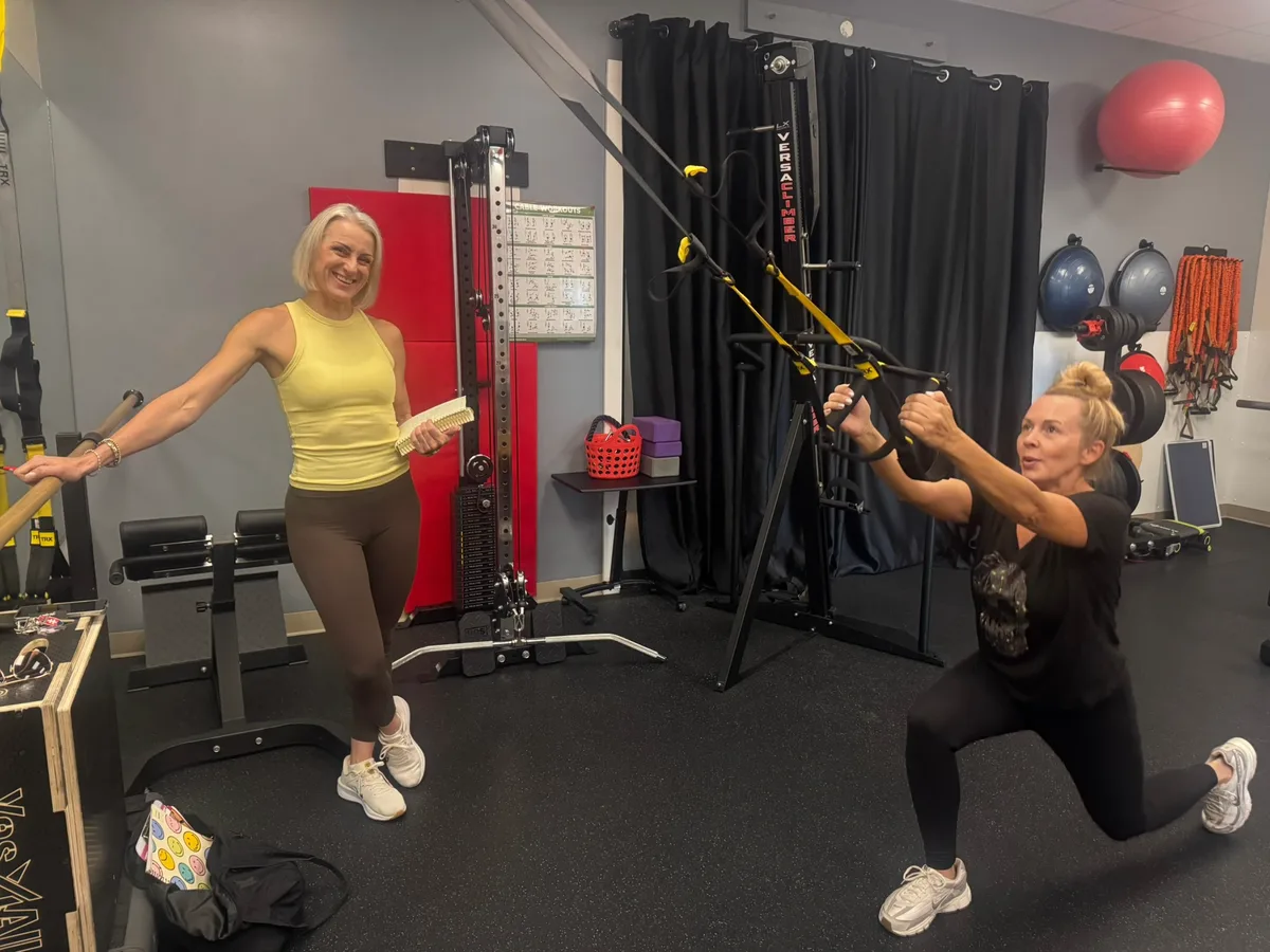 A trainer with a clipboard coaching a member through a TRX row in a lunge