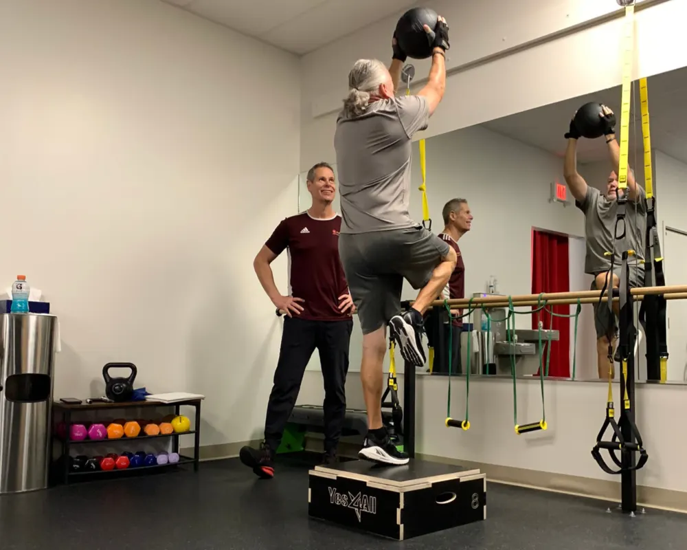 A trainer watching a member press a medicine ball overhead on a box