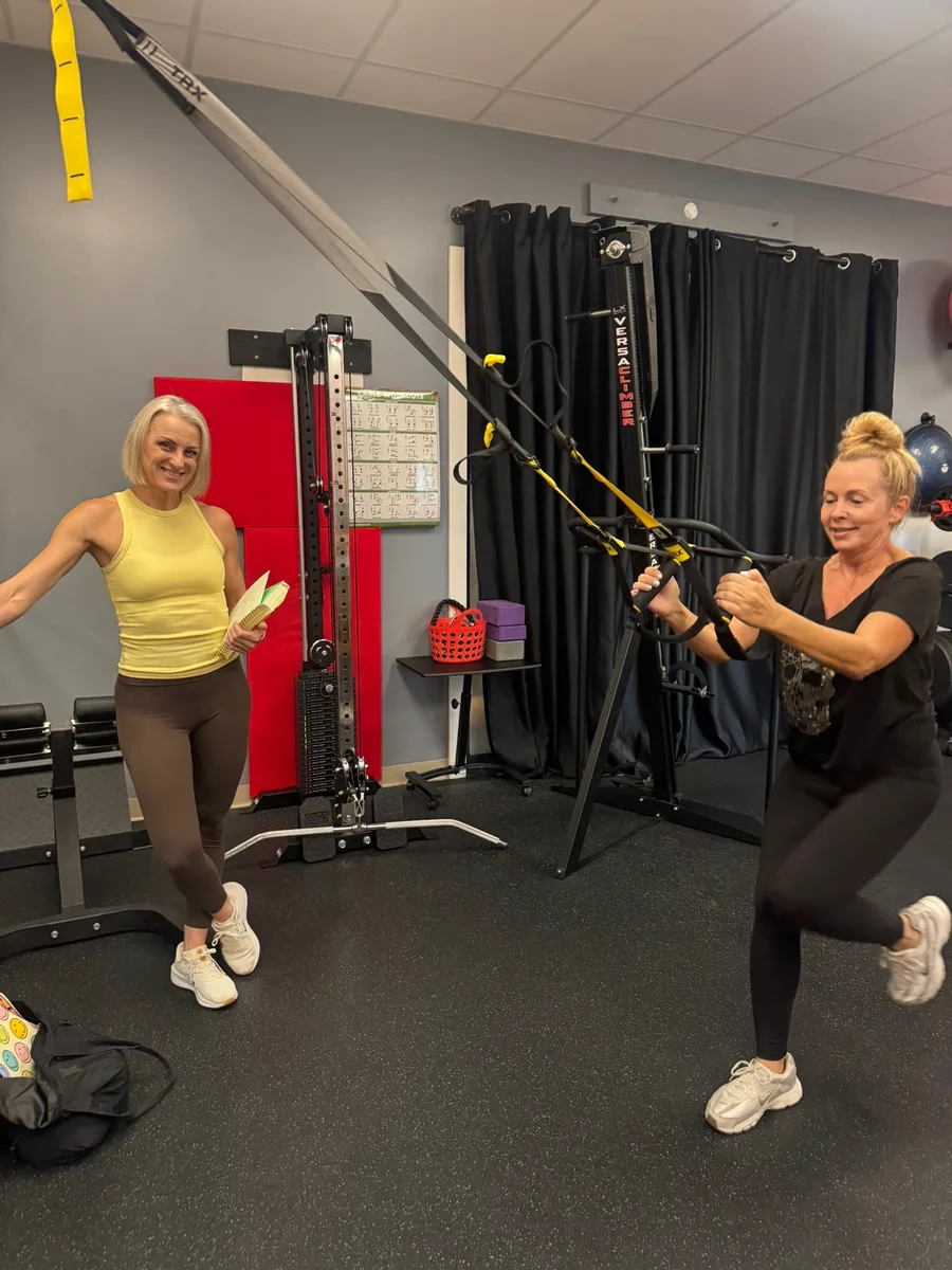 A trainer holding a clipboard beside a member doing a TRX row